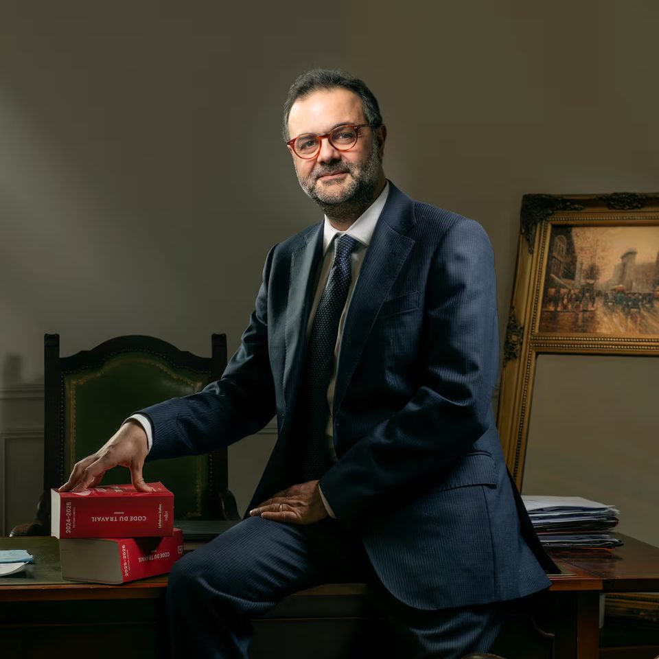 Portrait d’Avi Bitton, avocat au barreau de Paris, assis sur un bureau, la main posée sur deux éditions du Code du travail.